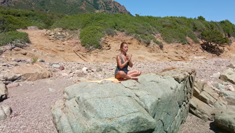 beautiful woman meditating with eyes closed and sitting on a rock by the sea in sardinia, italy - aerial pullback