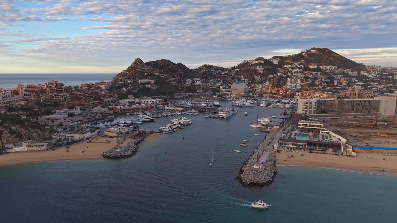 Et kredsende luftfoto, der viser bugten ved Cabo San Lucas, med dens luksuriøse marinaer og eksklusive yachter, der sejler langs kystlinjen ved solnedgang, Mexico