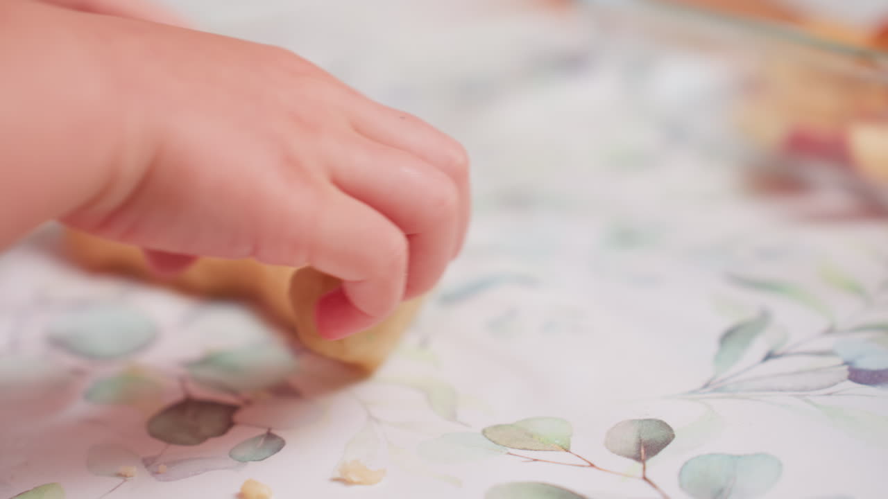 Hand view of toddler pressing dough gently on floral table mat during activity session, small fingers shaping mixture with focus while surrounded by kitchen items and play tools