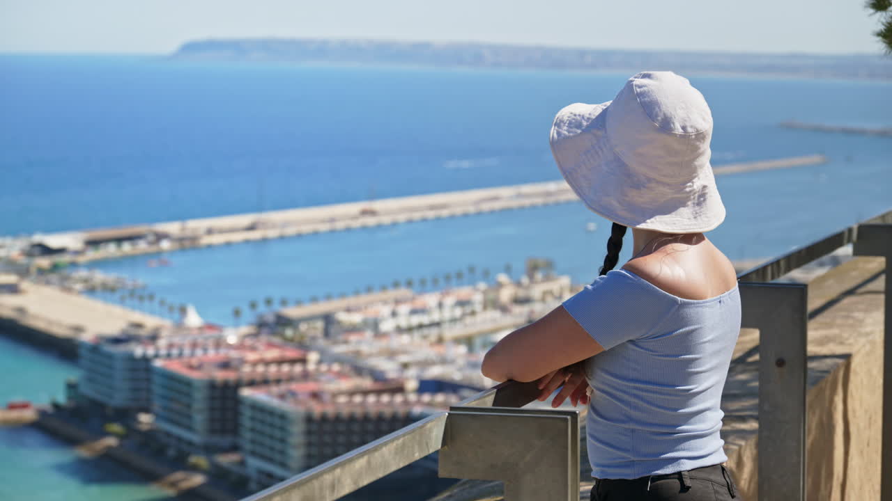 A traveler girl in a white bucket hat takes in the coast from a breezy hilltop lookout in Alicante, Spain