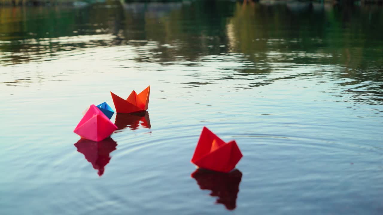 Woman's hand is putting coloured paper ships into the water in the evening. Bright origami boats in the river surface. Origami ship sailing.