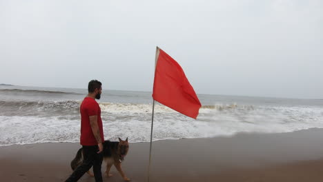 Perro-Pastor-Alemán-En-La-Playa-Jugando-Con-Su-Dueño-Cerca-De-La-Bandera-Roja