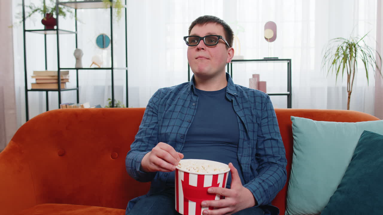 Caucasian man in 3d glasses sitting on couch eating popcorn snacks and watching interesting tv film