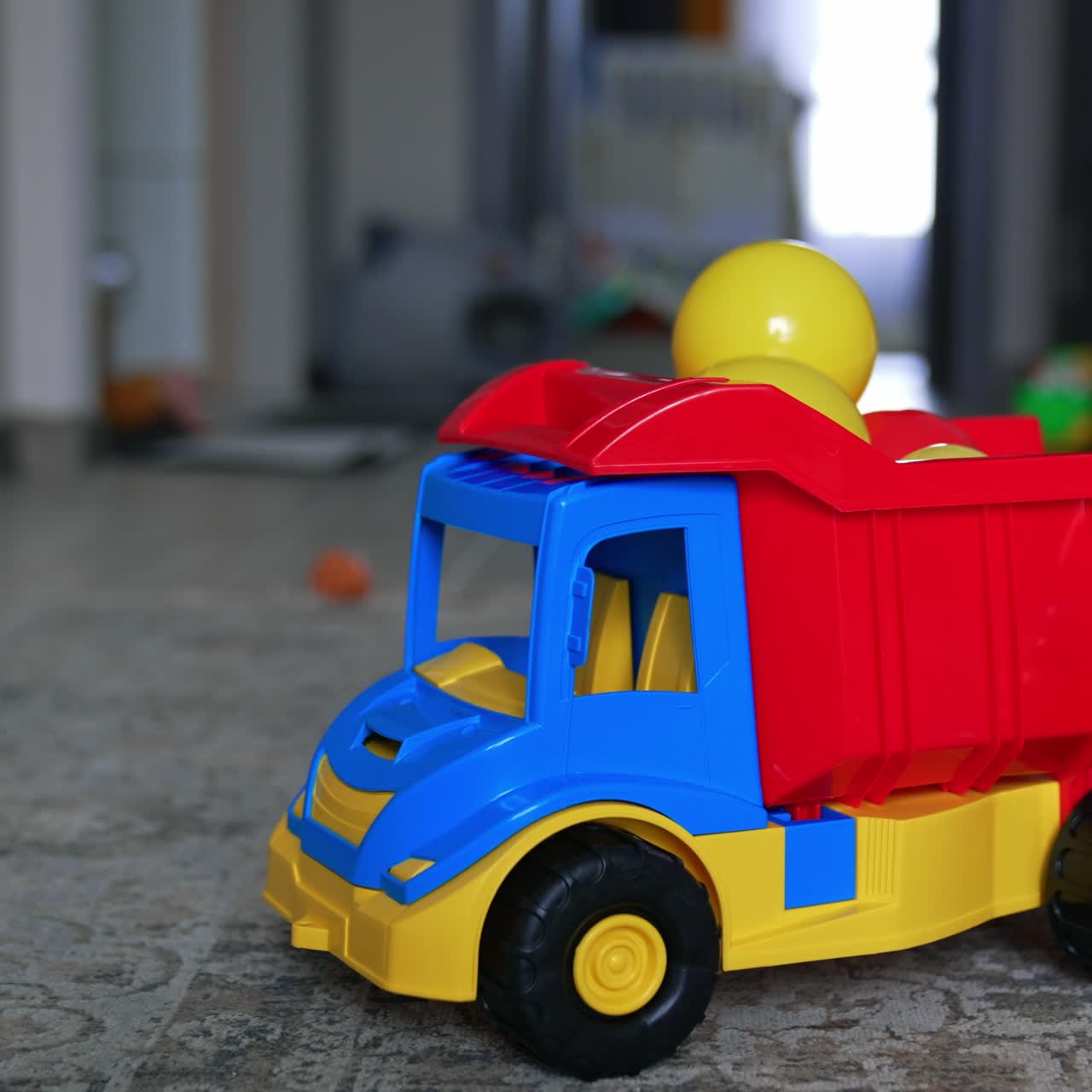 Bright toy lorry filled with plastic balls. Little kid in orange sport suit crawls out of the room. Blurred backdrop