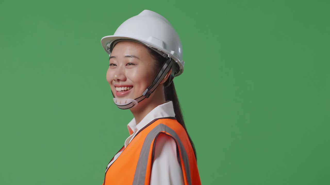 으로 가게 봤을 때, 안전 헬을 입은 아시아 여성 엔지니어가 녹색 스크린 배경 스튜디오에서 는 동안 주변을 둘러보고 있습니다.