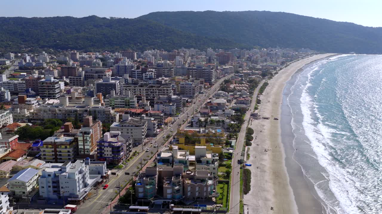 Bombinhas, State of Santa Catarina, Brazil, highlighting the urban development along the coastline, featuring buildings, roads, and the beach on a sunny day, drone orbiting