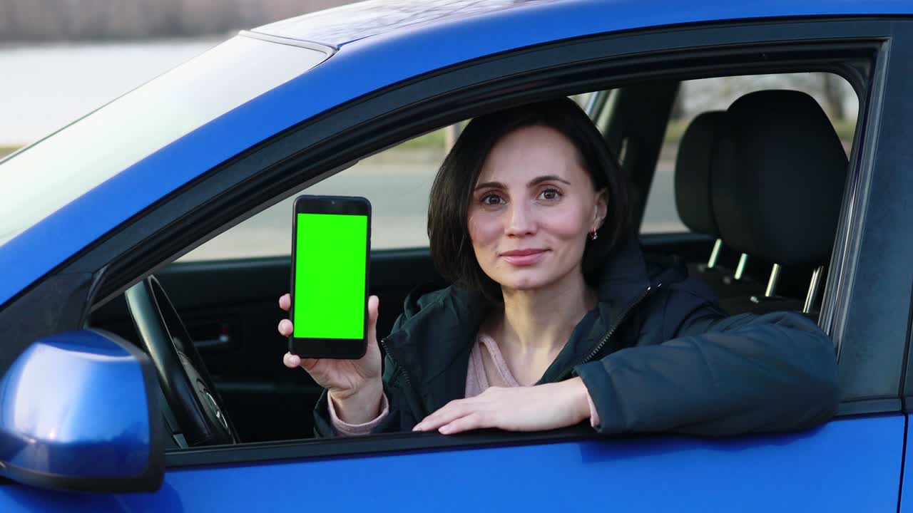 mujer europea con cabello corto sentada en el coche azul, sonriendo y mostrando un teléfono móvil con pantalla verde y su dedo índice está apuntando hacia el teléfono