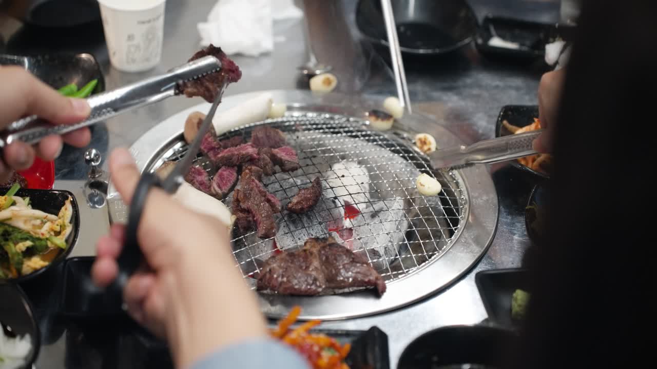 Hands with metal tongs and scissors slicing cuts of premium Korean Hanu beef steak over a hot charcoal grill surrounded by side dishes in a Korean barbecue restaurant