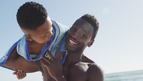 Portrait-of-smiling-african-american-father-carrying-his-son-on-sunny-beach