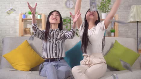 two happy asian girls catch flying dollar banknotes while sitting at home on the sofa in the living room