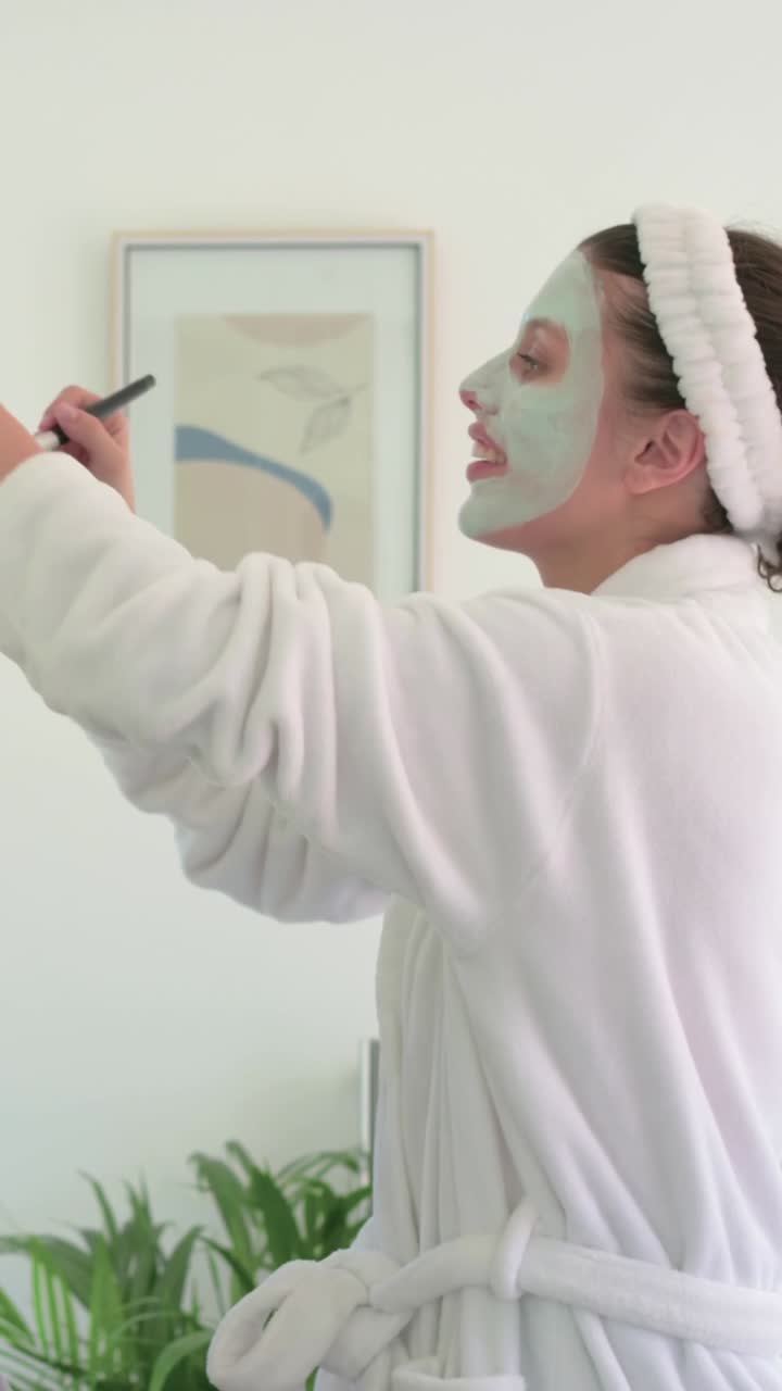 Video verticale: Solleva un oggetto scuro, donna in accappatoio bianco che posa a casa con una maschera verde, spazio per il testo