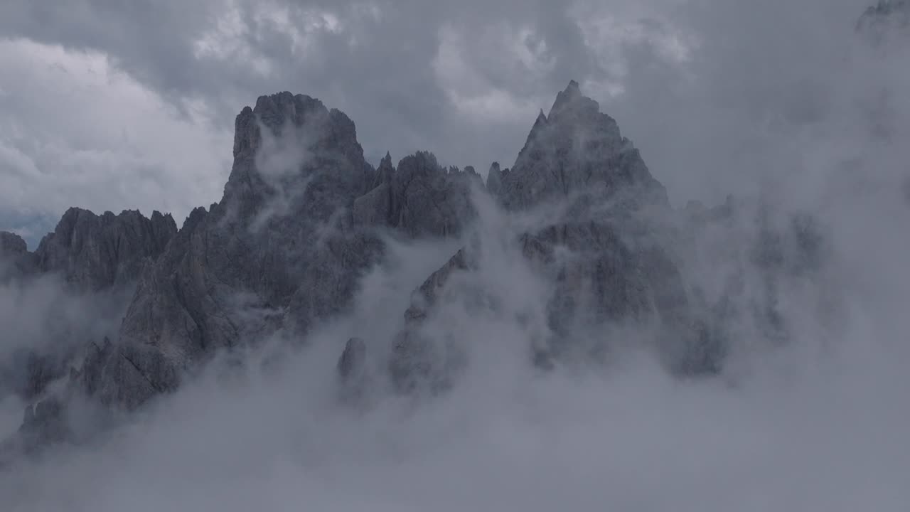 Majestic rocky mountain peaks emerge dramatically through swirling clouds and mist in moody overcast conditions. Jagged alpine summits pierce through thick fog creating mystical atmospheric landscape