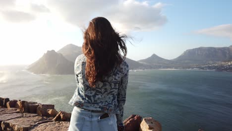 una hermosa bahía rodeada de montañas con una hermosa chica de pelo largo y oscuro mirando hacia las aguas mientras la cámara gira alrededor de ella en un arco