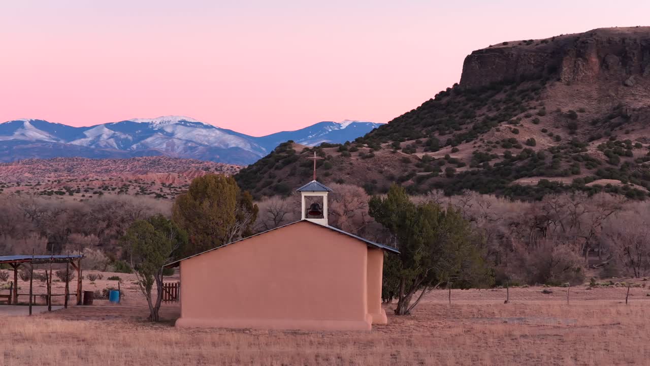 Un’orbita cinematografica di un drone a media distanza mostra una chiesa di campagna con croce e campanile al crepuscolo. La ripresa circola per rivelare un’enorme mesa e cime innevate. Perfetta per sequenze di apertura di documentari