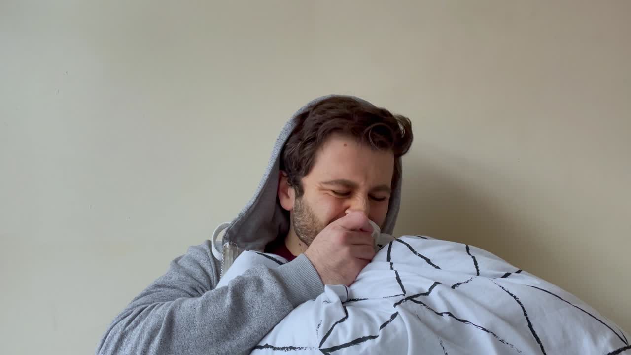 joven estudiante universitario masculino que parece visiblemente indispuesto tosiendo mientras habla con la cámara en una videollamada