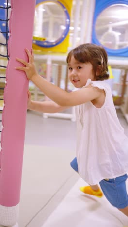 a happy and energetic 5-year-old girl laughs and smiles at the camera while running to push a colorful, soft merry-go-round at a modern indoor playground