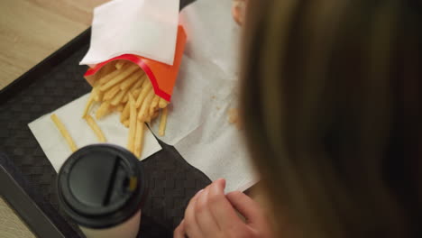 la mano de la mujer recoge suavemente una hamburguesa de una servilleta colocada en una bandeja, ya que las piezas de la hamburguesa han caído en la servilleta, la bandeja también contiene papas fritas y una taza de café colocada en la mesa de madera