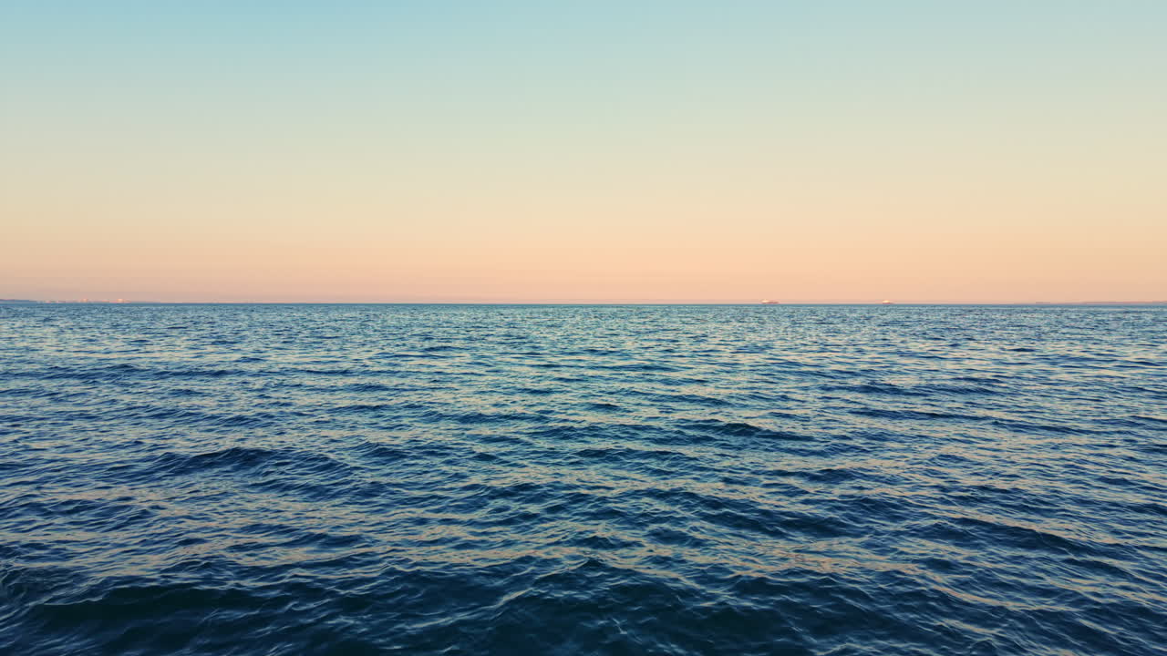 Waves move gently in the ocean as the sun sets on the horizon, creating a gradient of colors in the sky above the water