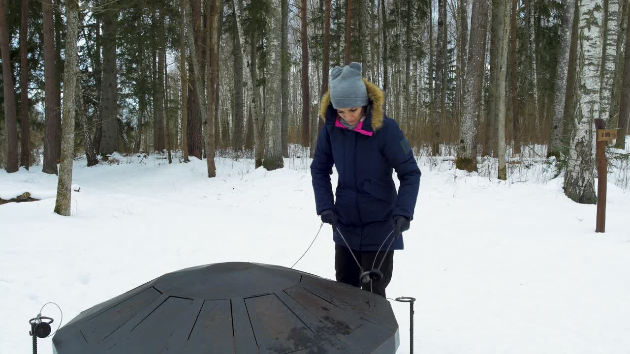 En ung kaukasisk flicka spelar på en stor steeldrum i en snötäckt vintertallskog en solig dag, ensam i frostigt väder, inzoomande gimbal-tagning