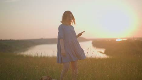 woman in blue gown enjoys a tranquil sunset by the lake, smartphone in hand, with hair gently swaying in the breeze