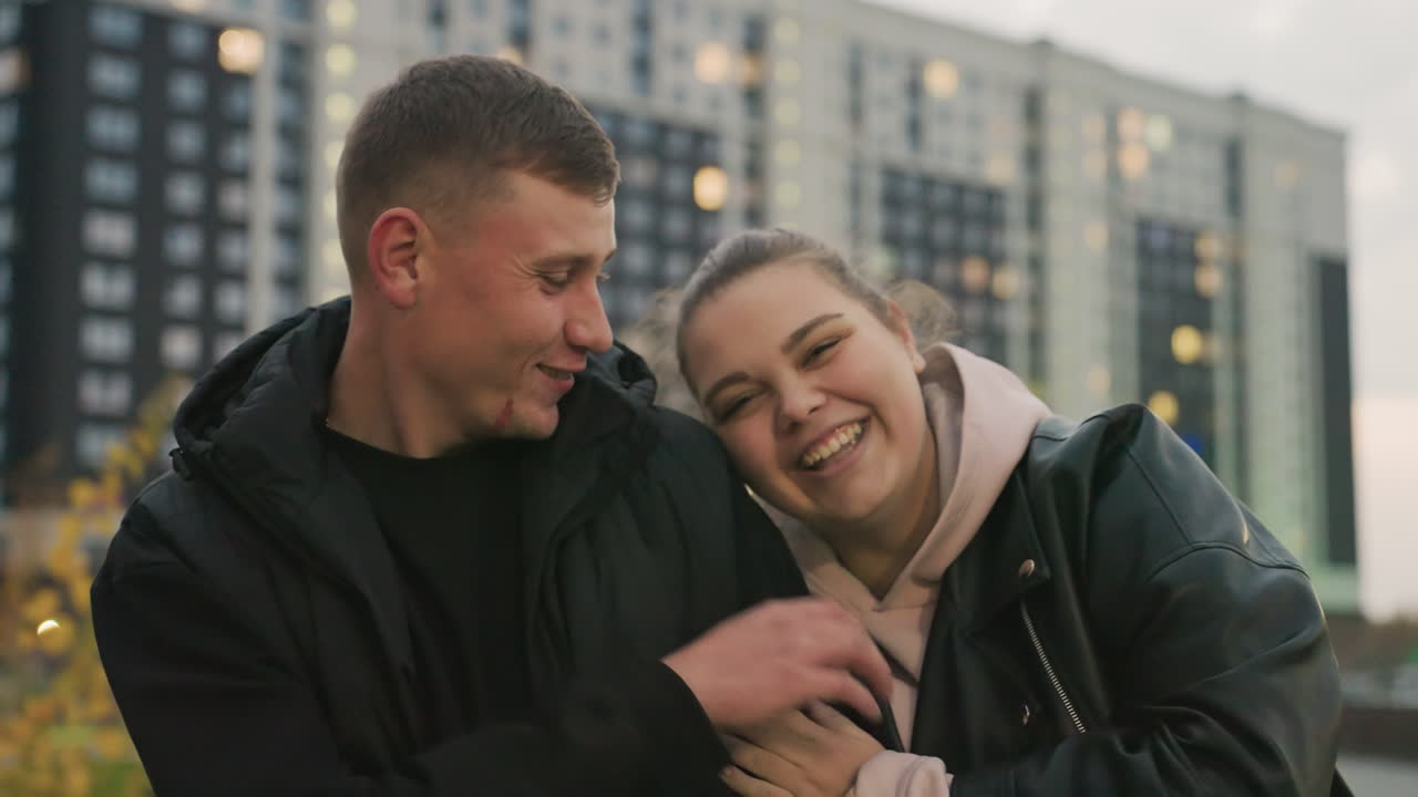 Lady rests head on her lover shoulder while gently tickling his ear with soft playful gesture as they share romantic connection smiling together during calm evening walk near city buildings