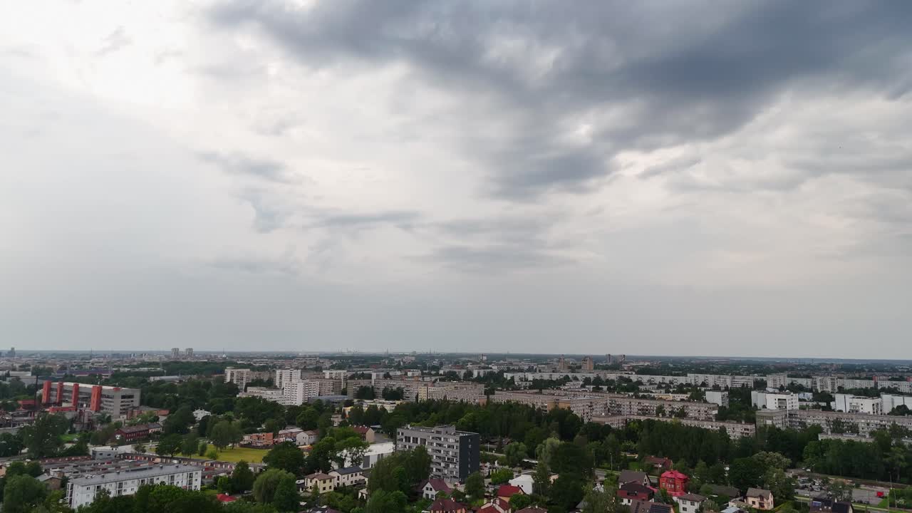The drone tilts down over Riga’s Darzciems district, revealing a mix of small houses, trees, and distant modern buildings under a cloudy sky