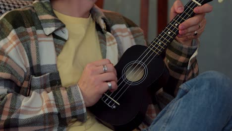 primer plano de un tipo con cabello moreno rizado en una camisa a cuadros tocando el ukulele con su mano con dos anillos en un apartamento moderno