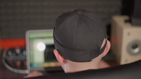 a man's head wearing a black cap, sitting on a black chair, working on a computer. the camera angle is from behind, showing the man focused on his task as the camera zooms in