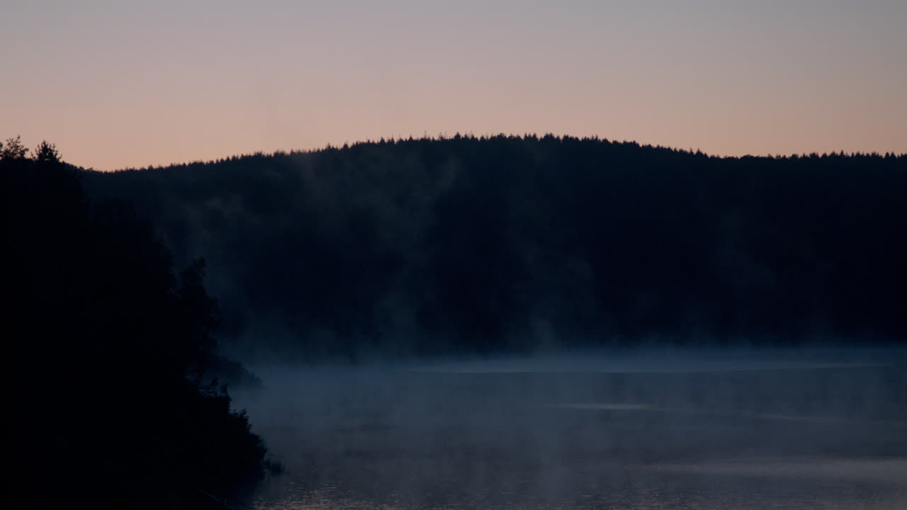 High quality video shows a calm morning lake wrapped in soft mist with gentle sunlight rising behind a dark forest line creating a serene and atmospheric nature moment