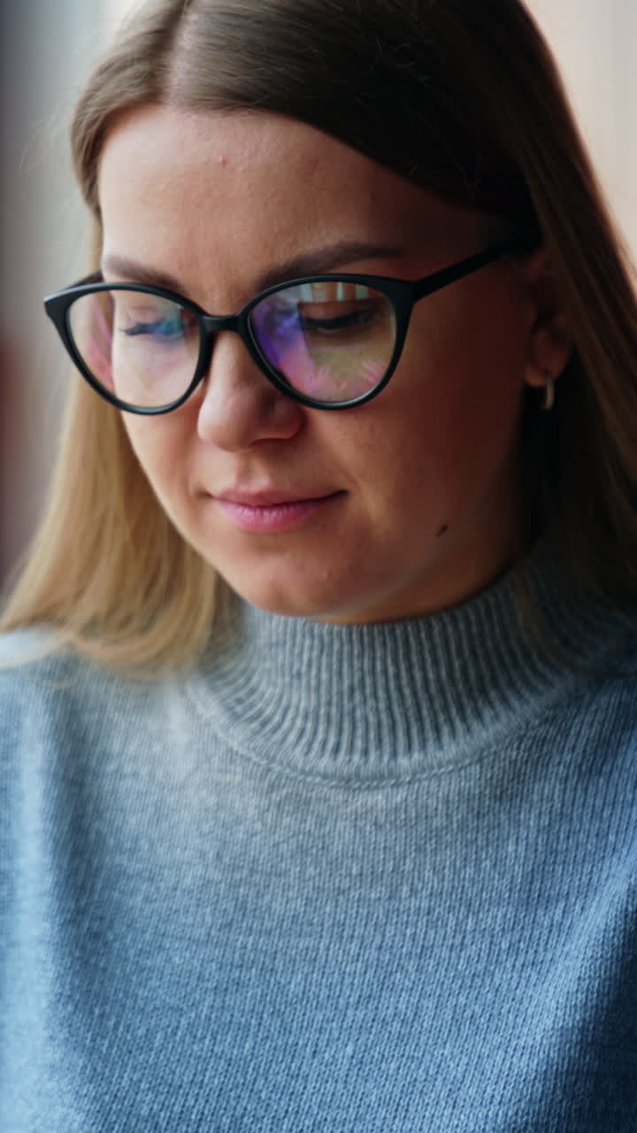 Blonde lady wearing glasses is focused on her work on laptop. Woman is typing something, then looks at camera and smiles. Blurred backdrop. Vertical video