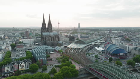 Drone-Filma-En-Cámara-Lenta-La-Catedral-De-Colonia,-La-Estación-Central-De-Colonia,-El-Teatro-De-Colonia-En-La-Puesta-Del-Sol,-El-Drone-Se-Mueve-Hacia-Abajo-Y-El-Rin-Con-El-Puente-Hohenzoller-Entra-En-Escena