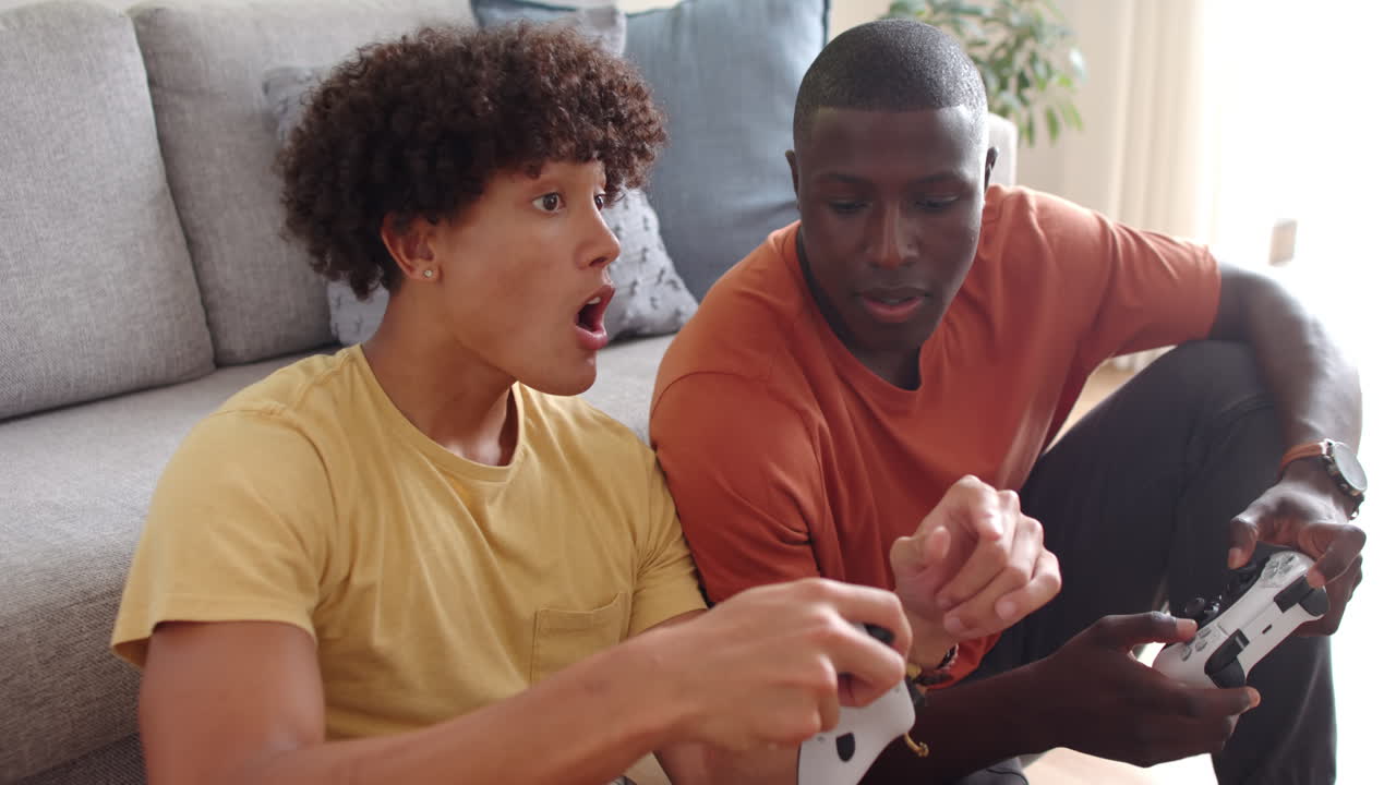 Playing video games, two multiracial male friends sitting on floor, enjoying leisure time, at home
