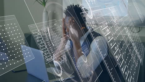 man pressing temples, leaning over laptop, viewing animated finance graphs and data overlays