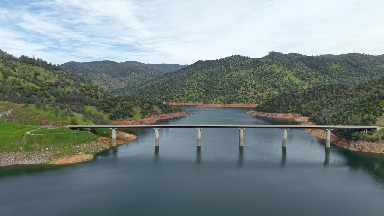 This ascending high-angle drone shot offers a dynamic view of the Don Pedro Reservoir Bridge, showing the contrast between the smooth water surface and the rugged terrain.