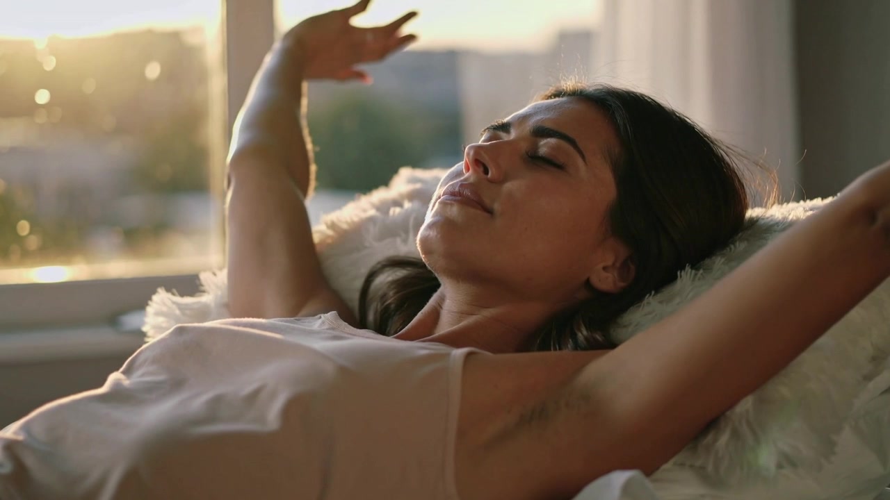 Donna in canottiera che alza le braccia e si allunga sul letto con un cuscino vicino alla finestra, a causa della luce del sole.