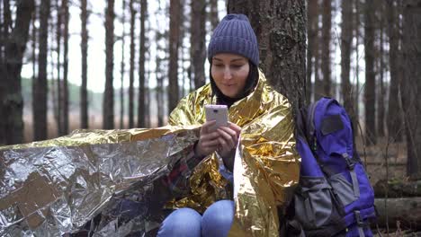 positive female tourist wrapped in a golden forest survival blanket uses a smartphone