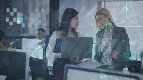 Animation-of-globes-of-digital-icons-over-two-diverse-women-discussing-over-a-laptop-at-office