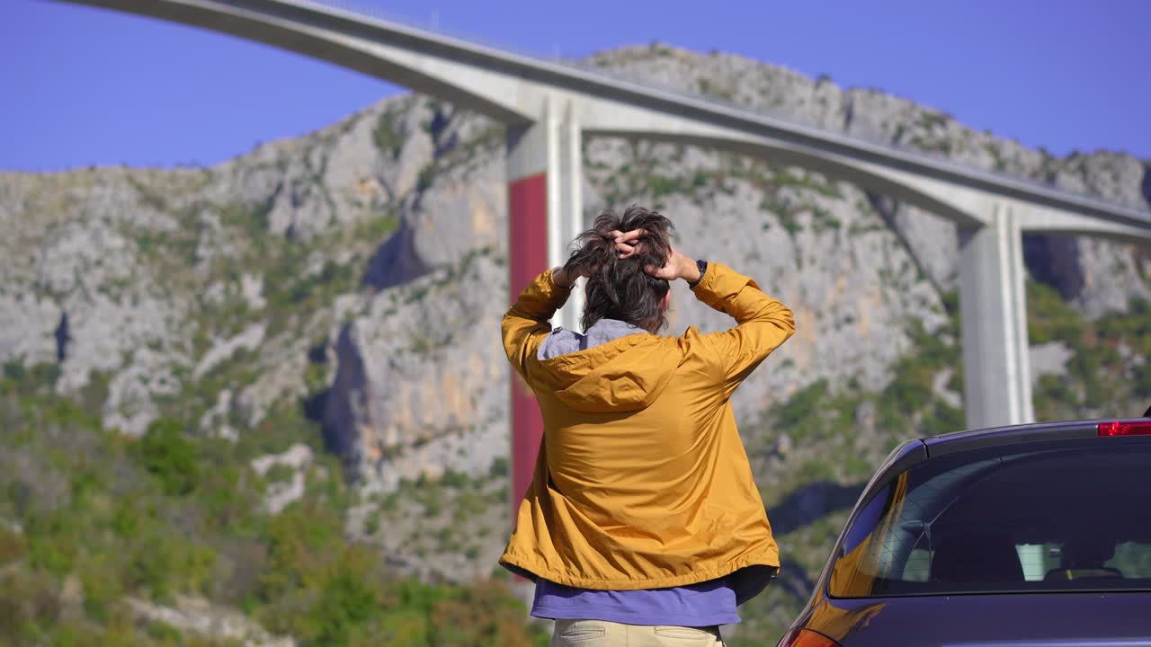 un hombre de pie junto a su coche mira el puente de moracica en montenegro. está conmocionado por las noticias de lo caro que es este puente y cuánto dinero le debe ahora montenegro a china.