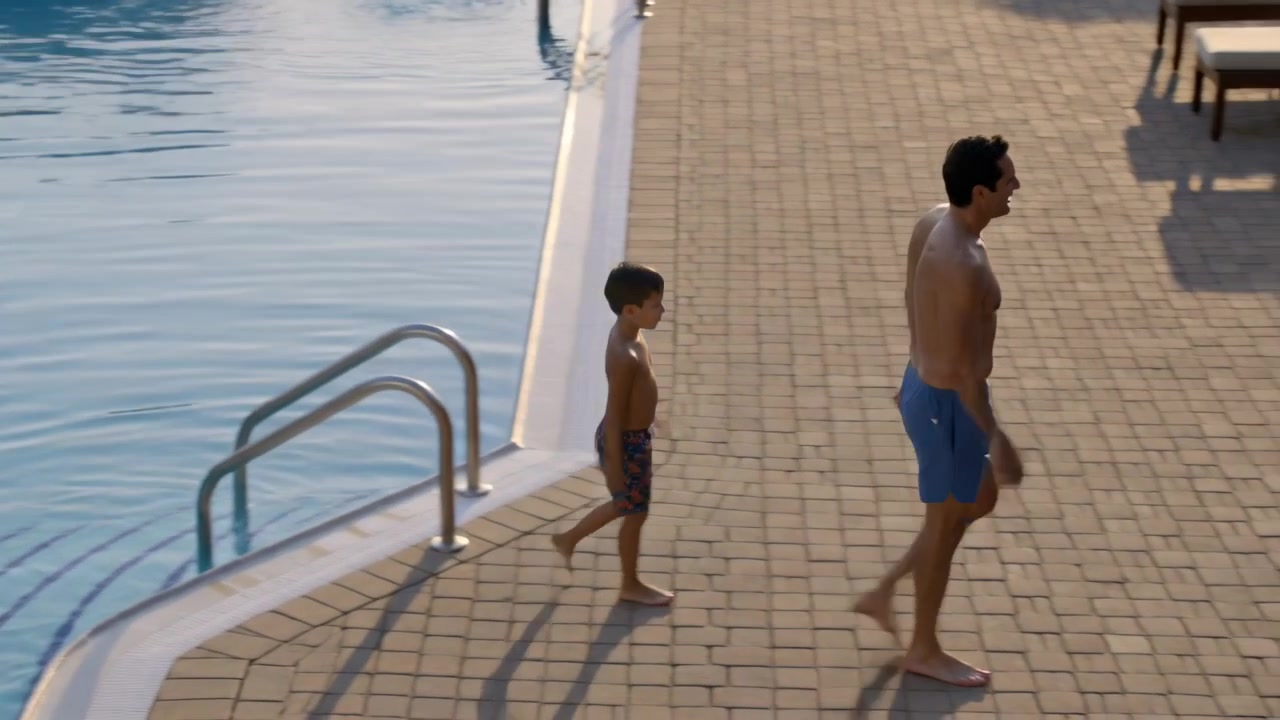 Testing pool water, dad in trunks and son walking from ladder toward lounge chairs, copy space