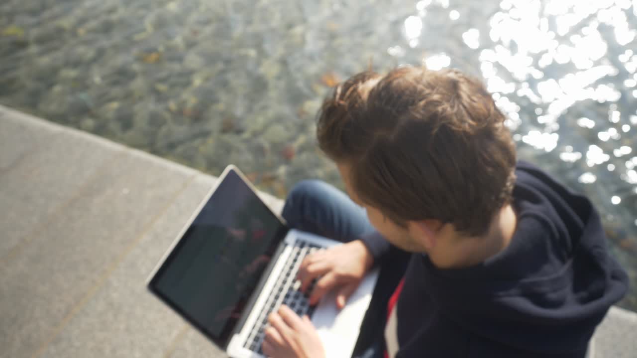 Focus pull of a young man sitting on the bank of a canal in Amsterdam typing on the notebook