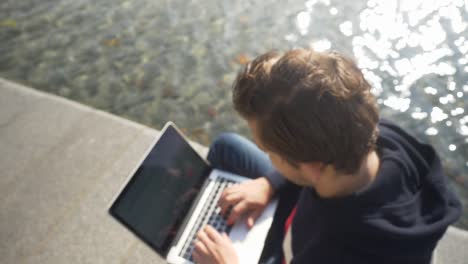 Focus-pull-of-a-young-man-sitting-on-the-bank-of-a-canal-in-Amsterdam-typing-on-the-notebook