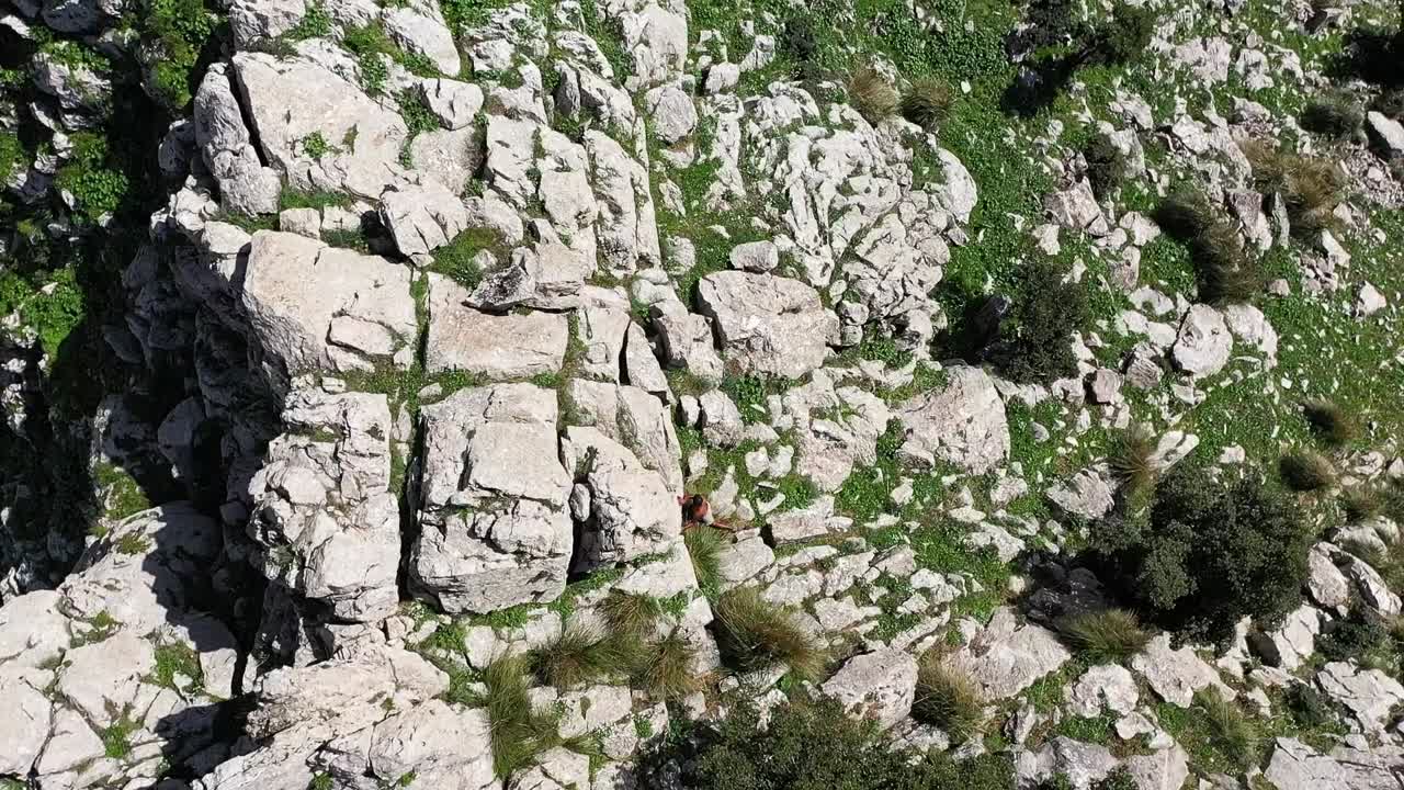 혹독한 햇빛이 내리쬐는 더운 여름날 튀니지의 자구안 정상까지 오르기 위해 바위 사이를 걸어가는 현지 흑인의 회전하는 드론 샷