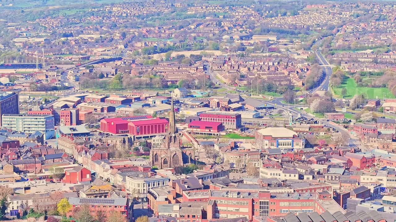 Aerial ascending view of Rotherham town centre showing the All Saints Church spire rising above red‑roofed historic buildings, civic blocks, retail frontage, and surrounding residential streets