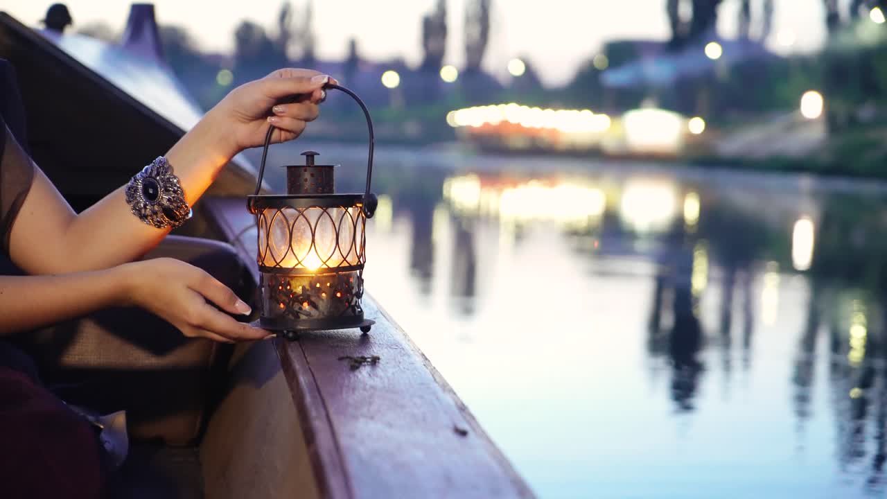 hands of a girl are holding a decorative lantern on a boat in a river near the bank