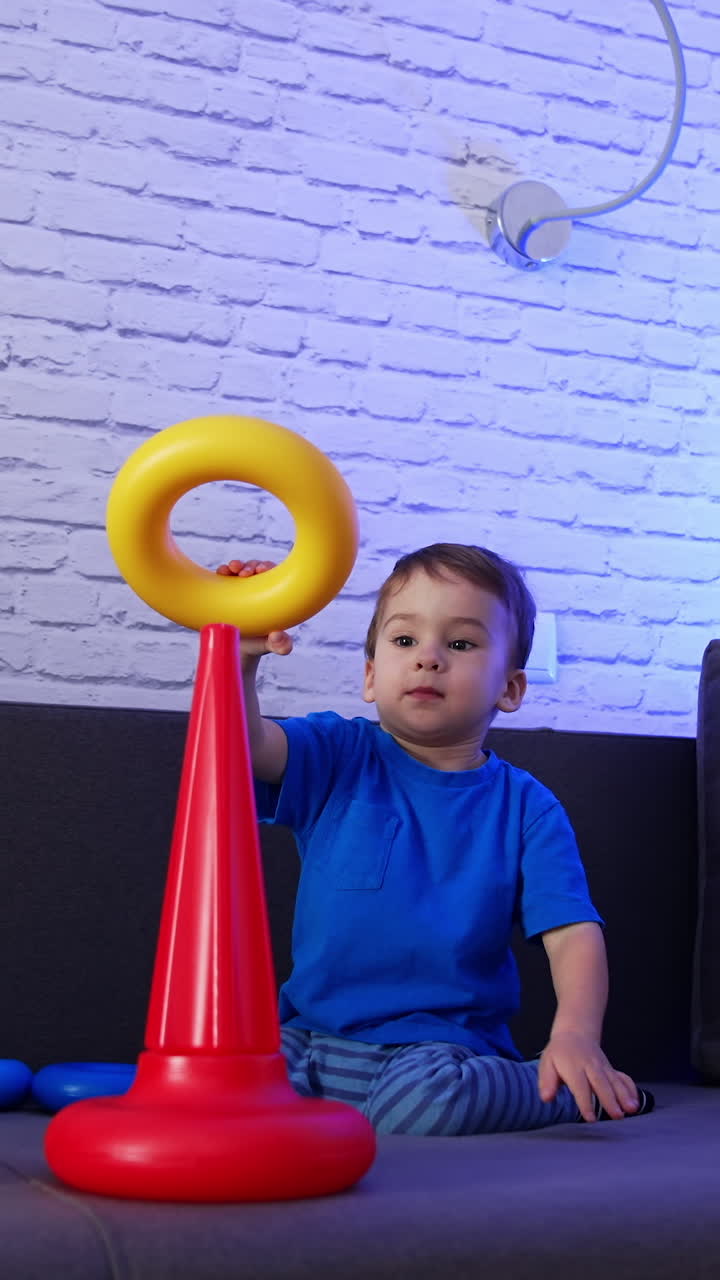 Cute dark-haired toddler assembling pyramid on the sofa. Baby boy plays with toy at home. Vertical video