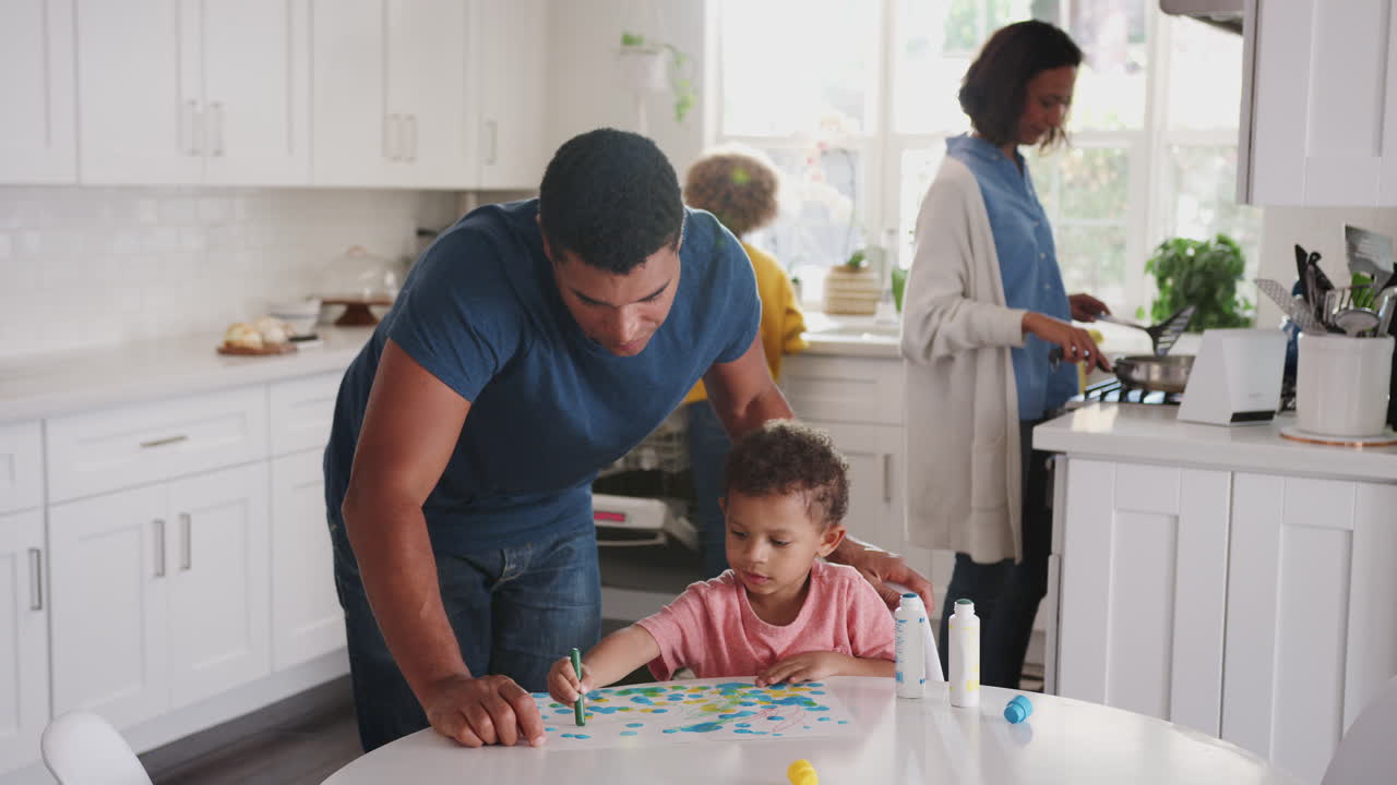 Dad watches son painting in kitchen, returns to chores with mother and daughter in the background