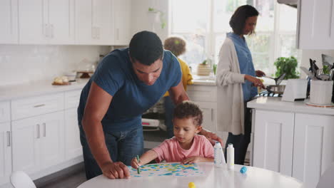 Papá-Mira-A-Su-Hijo-Pintando-En-La-Cocina,-Vuelve-A-Las-Tareas-Del-Hogar-Con-Madre-E-Hija-En-El-Fondo