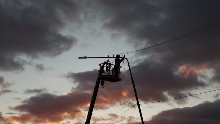 A silhouetted worker in a cherry picker adjusts overhead power lines against a twilight sky with orange-tinged clouds, remaining stationary on the elevated platform as the sky slowly shifts.