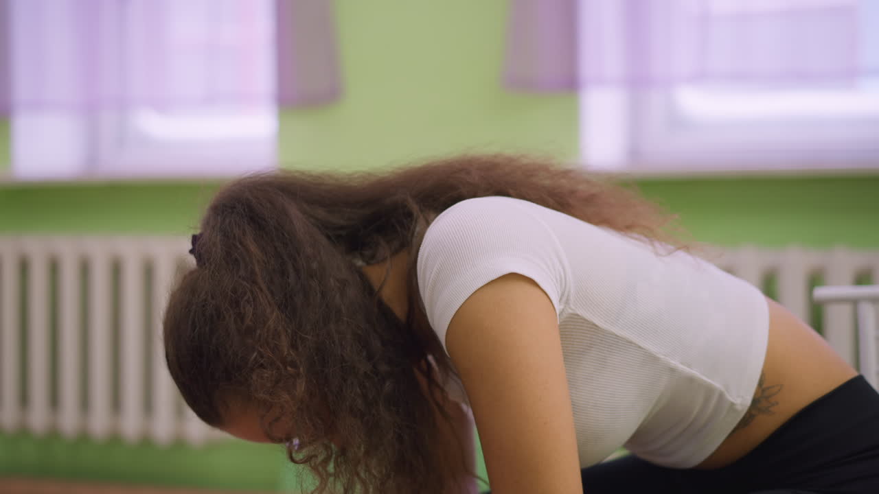 Closeup view of young woman in white top and black leggings with glasses sitting on chair resting hands on knees looking calm and relaxed inside studio with green wall and blurred workout