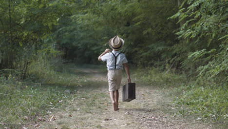 boy wearing vintage clothes and a hat walking away from camera barefoot in the forest holding a stick and a suitcase in summer